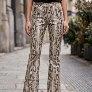 May include: High-waisted trousers featuring a beige and dark brown snakeskin print pattern. These trousers have a straight-leg cut with a slight flare at the hem, a button closure at the waist, and visible belt loops. The material has a smooth texture.