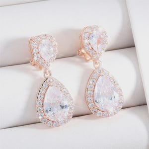 Peut inclure: Une paire de boucles d'oreilles pendantes en or rose avec une pierre claire en forme de poire à la base et une pierre claire plus petite en forme de poire au sommet, entourées d'un halo de minuscules pierres claires rondes.
