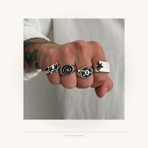 Peut inclure: Quatre bagues argentées portées sur les doigts. La première présente deux étoiles noires, la deuxième une spirale noire, la troisième une étoile noire avec un cercle central, et la quatrième une face rectangulaire avec une étoile noire. Le texte VELURO APT - Handcrafted Jewelry est visible.