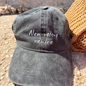 Può includere: Un cappellino da baseball grigio antracite con una finitura in tessuto slavato e vissuto. Il pannello frontale presenta una scritta ricamata in bianco che recita "Non volevo venire" in un semplice carattere corsivo. Il cappello ha una visiera curva.