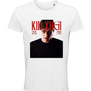 Può includere: Una t-shirt bianca a maniche corte con una stampa grafica centrale che ritrae una persona con una maglia nera. Il testo KID YUGI appare in rosso sopra il ritratto, con ANCHE QUESTO È MILANO e SUMMER TOUR 2024 stampati in rosso ai lati.