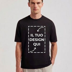 Op de afbeelding: Een zwart t-shirt met korte mouwen en een wit gestippeld rechthoekig kader op de borst. Binnen het kader staat de tekst IL TUO DESIGN QUI, vergezeld door twee witte pijlen die naar het midden van het grafische gedeelte wijzen.
