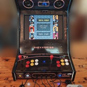 Puede incluir: Una máquina recreativa negra con una pantalla que muestra el menú del juego Mortal Kombat, dos joysticks rojos, botones de control coloridos y cuatro mandos de juego grises con cable conectados al panel frontal con el texto Quack Station.