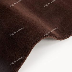 May include: A solid dark brown rug with a soft, dense pile texture. The surface features the repeating white text 'Rug Aura Homes' printed across the fabric. The rug is shown folded, displaying its thick, plush construction and deep brown color.