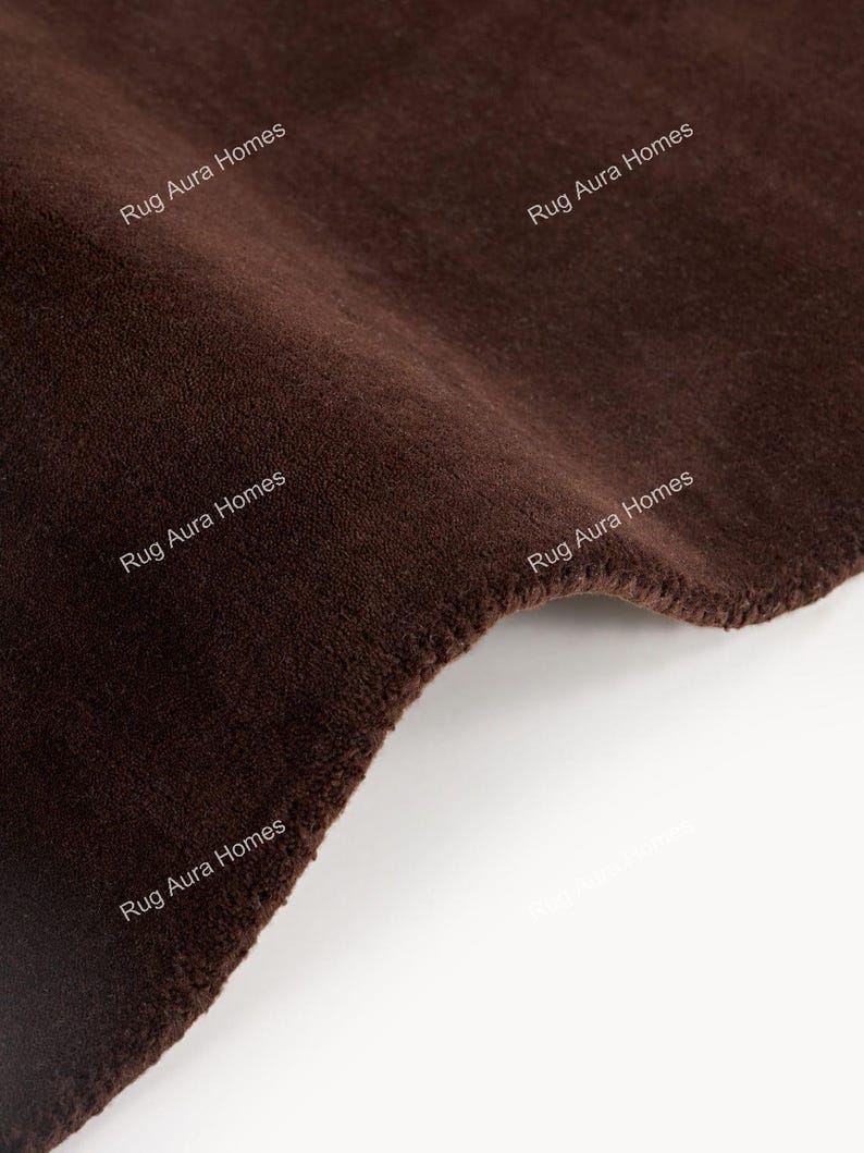 May include: A solid dark brown rug with a soft, dense pile texture. The surface features the repeating white text 'Rug Aura Homes' printed across the fabric. The rug is shown folded, displaying its thick, plush construction and deep brown color.