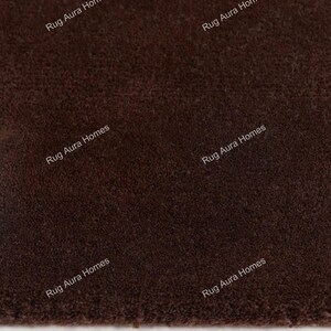 May include: A solid dark brown rug with a dense, short-pile texture. The surface features the repeating white text "Rug Aura Homes" printed across the fabric in a diagonal orientation, indicating the brand name on the textile material.