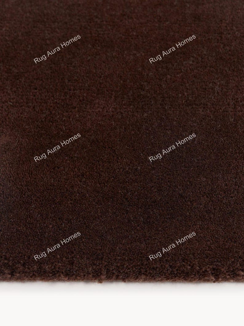 May include: A solid dark brown rug with a dense, short-pile texture. The surface features the repeating white text "Rug Aura Homes" printed across the fabric in a diagonal orientation, indicating the brand name on the textile material.