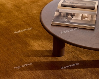 May include: A solid mustard-gold area rug with a soft, uniform texture. A dark brown circular wooden coffee table sits on the rug, supporting an open book with black and white interior photography. The text RugAuraHomes appears as a watermark.