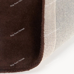 May include: A dark brown rug with a soft, solid-colored surface and a folded corner revealing a light-colored, woven backing with a herringbone pattern. The text Rug Aura Homes is printed multiple times across the surface of the rug.