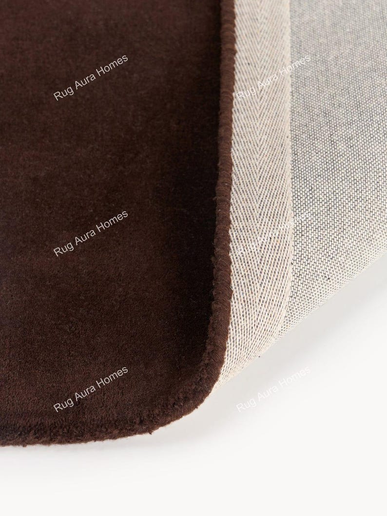 May include: A dark brown rug with a soft, solid-colored surface and a folded corner revealing a light-colored, woven backing with a herringbone pattern. The text Rug Aura Homes is printed multiple times across the surface of the rug.