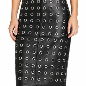 May include: A black leather pencil skirt featuring a high-waisted design and a vertical seam down the center. The entire surface is adorned with a repeating grid pattern of silver-toned metal grommets, creating a structured and uniform aesthetic.