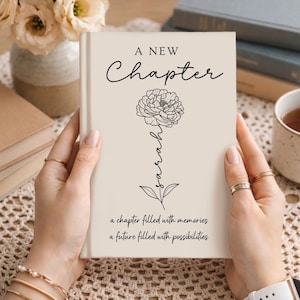 May include: A beige hardcover book featuring the text A NEW Chapter at the top, a black line art illustration of a flower with the name Sarah written vertically along the stem, and the text a chapter filled with memories a future filled with possibilities.