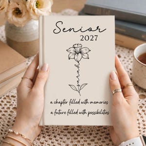 May include: A beige hardcover journal featuring the text Senior 2027 at the top, a black line art illustration of a daffodil with the name Sarah written vertically along the stem, and the phrase a chapter filled with memories a future filled with possibilities.