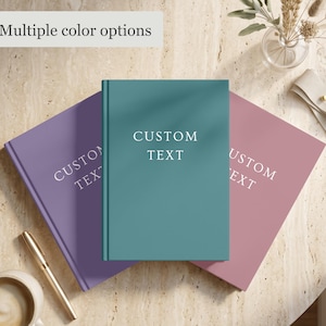May include: Three hardcover journals in purple, teal, and dusty rose, each featuring the white printed text CUSTOM TEXT on the front cover. The journals are arranged in a fan shape, showcasing the different color options available for customization.