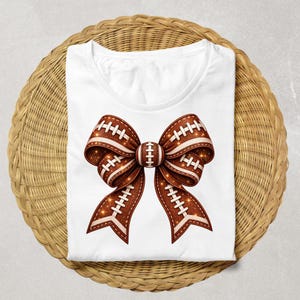 Puede incluir: Una camiseta blanca con un gráfico central grande de un lazo decorativo. El lazo es de color marrón con una apariencia texturizada y brillante, decorado con cordones blancos que imitan el diseño de un balón de fútbol americano.