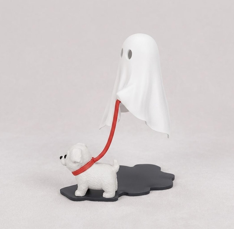 Puede incluir: Una figura de fantasma blanco de pie, conectada por una correa roja a una figura de cachorro blanco. Ambas figuras est&aacute;n montadas sobre una base negra con forma de nube. El fantasma tiene dos orificios oculares circulares y el cachorro ojos negros.
