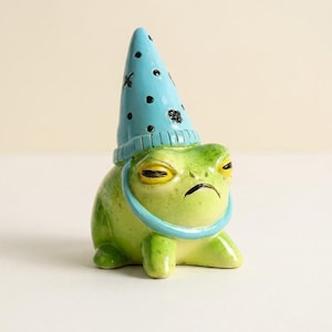 Könnte beinhalten: Eine Keramikfrosch-Figur mit glänzend grüner Oberfläche und gelben Augen. Der Frosch trägt einen hellblauen, spitzen Hut mit schwarzen Punkten und kleinen schwarzen Insektenabbildungen sowie ein passendes blaues Band um den Halsbereich.