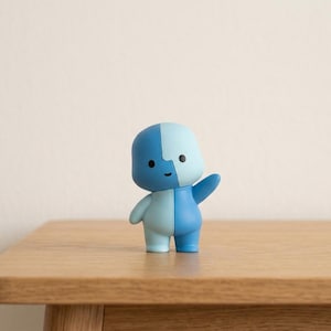 Può includere: Una piccola statuetta stilizzata con testa e corpo arrotondati, divisi verticalmente in due distinte tonalità di blu. Il personaggio presenta due semplici occhi a punto nero, una piccola bocca curva e un braccio sollevato in un gesto di saluto.