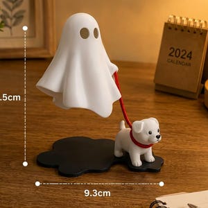 Puede incluir: Una figura de fantasma blanco que sostiene una correa roja unida a una peque&ntilde;a figura de cachorro blanco. El fantasma y el cachorro se apoyan sobre una base negra. El objeto mide 11,5 cm de alto y 9,3 cm de ancho seg&uacute;n las l&iacute;neas de medida.