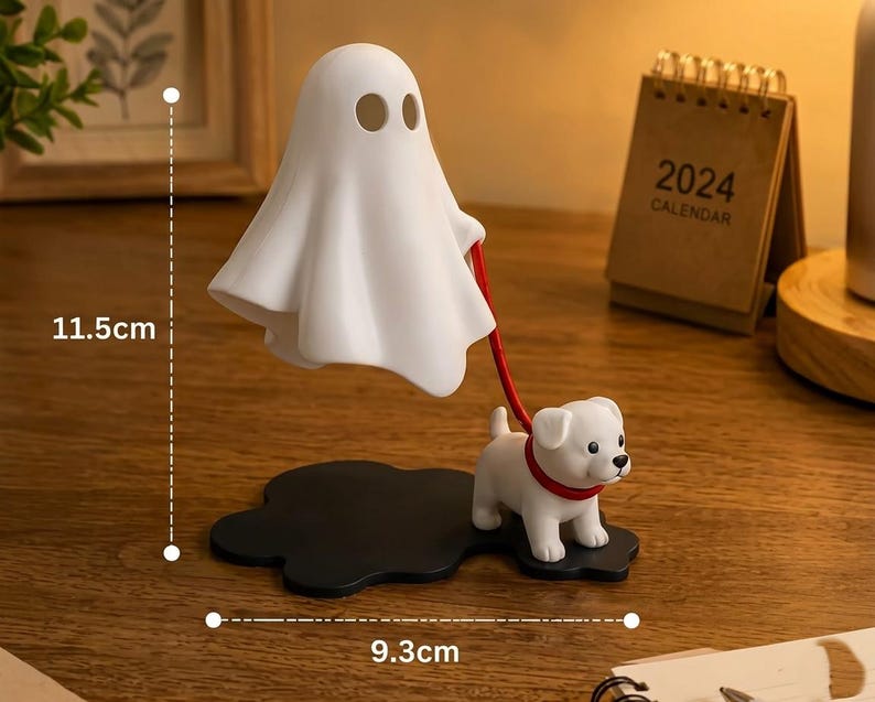 Puede incluir: Una figura de fantasma blanco que sostiene una correa roja unida a una peque&ntilde;a figura de cachorro blanco. El fantasma y el cachorro se apoyan sobre una base negra. El objeto mide 11,5 cm de alto y 9,3 cm de ancho seg&uacute;n las l&iacute;neas de medida.