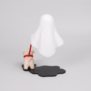Puede incluir: Una l&aacute;mpara decorativa que presenta una peque&ntilde;a figura de cachorro de color claro sobre una base negra con forma de charco. El cachorro lleva un collar rojo unido a una pantalla blanca con forma de fantasma que flota sobre la figura.
