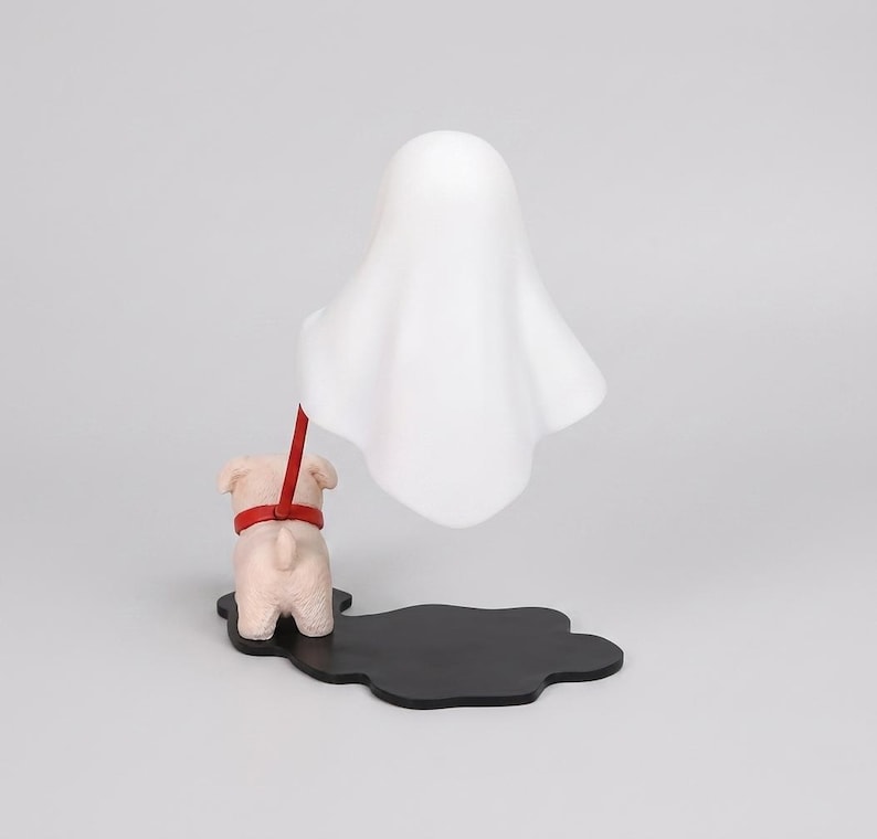 Puede incluir: Una l&aacute;mpara decorativa que presenta una peque&ntilde;a figura de cachorro de color claro sobre una base negra con forma de charco. El cachorro lleva un collar rojo unido a una pantalla blanca con forma de fantasma que flota sobre la figura.