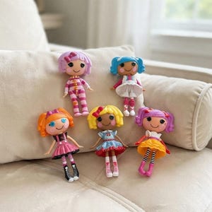 Peut inclure: Cinq petites poupées en plastique avec de grands yeux en forme de boutons et des cheveux en laine. Les poupées portent des tenues colorées, notamment un pyjama rayé, une robe blanche, une robe rose avec des bottes noires et une jupe jaune.