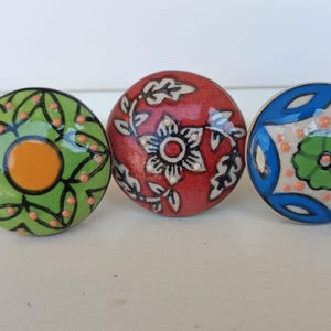Peut inclure: Trois boutons ronds en c&eacute;ramique avec des motifs peints &agrave; la main. Le bouton de gauche est vert avec un centre orange et des lignes noires. Le bouton central est rouge avec un motif floral blanc. Le bouton de droite est bleu et blanc.