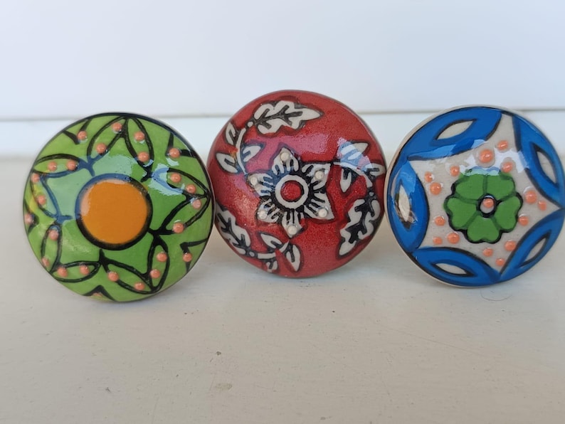 Peut inclure: Trois boutons ronds en c&eacute;ramique avec des motifs peints &agrave; la main. Le bouton de gauche est vert avec un centre orange et des lignes noires. Le bouton central est rouge avec un motif floral blanc. Le bouton de droite est bleu et blanc.