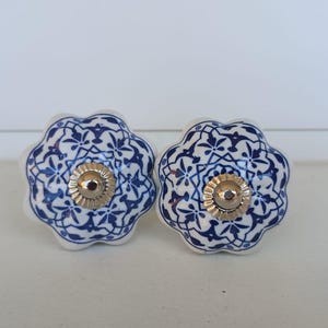 Peut inclure: Deux boutons de tiroir en c&eacute;ramique avec une forme festonn&eacute;e rappelant une fleur. Chaque bouton pr&eacute;sente une base blanche avec un motif floral et g&eacute;om&eacute;trique bleu d&eacute;taill&eacute;. Un capuchon en m&eacute;tal argent&eacute; est centr&eacute; sur le devant.