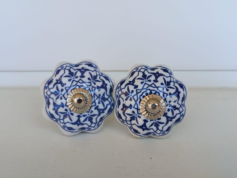 Peut inclure: Deux boutons de tiroir en c&eacute;ramique avec une forme festonn&eacute;e rappelant une fleur. Chaque bouton pr&eacute;sente une base blanche avec un motif floral et g&eacute;om&eacute;trique bleu d&eacute;taill&eacute;. Un capuchon en m&eacute;tal argent&eacute; est centr&eacute; sur le devant.