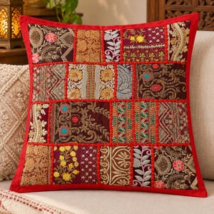 May include: A square throw pillow cover featuring a patchwork design of various embroidered fabric panels in shades of red, gold, brown, and green. The panels are separated by red fabric borders and decorated with floral and geometric patterns.