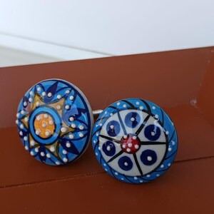 Peut inclure: Deux boutons de placard ronds en c&eacute;ramique avec des motifs peints &agrave; la main. Le bouton de gauche pr&eacute;sente un centre jaune avec des p&eacute;tales en forme d'&eacute;toile bleus et des points blancs en relief. Celui de droite a un centre rouge.