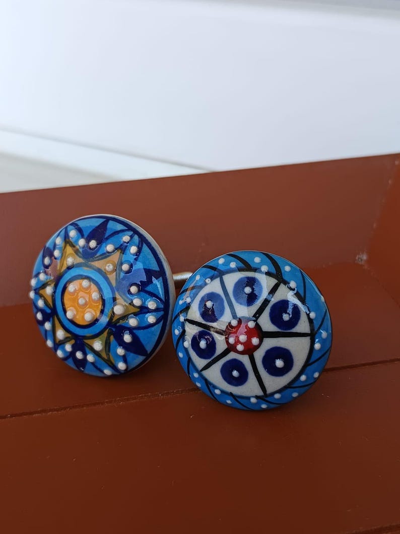 Peut inclure: Deux boutons de placard ronds en c&eacute;ramique avec des motifs peints &agrave; la main. Le bouton de gauche pr&eacute;sente un centre jaune avec des p&eacute;tales en forme d'&eacute;toile bleus et des points blancs en relief. Celui de droite a un centre rouge.