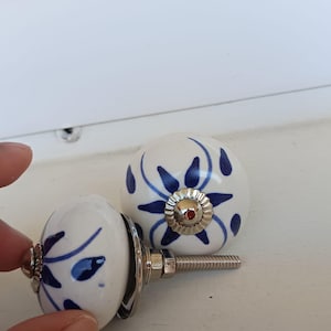 Peut inclure: Deux boutons de placard ronds en c&eacute;ramique avec une base blanche et des motifs floraux bleus peints &agrave; la main. Chaque bouton poss&egrave;de un accent central en m&eacute;tal argent&eacute; et une tige filet&eacute;e m&eacute;tallique &agrave; l'arri&egrave;re pour la fixation.