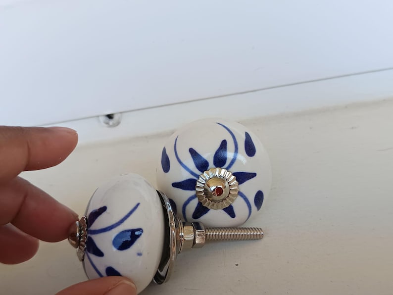 Peut inclure: Deux boutons de placard ronds en c&eacute;ramique avec une base blanche et des motifs floraux bleus peints &agrave; la main. Chaque bouton poss&egrave;de un accent central en m&eacute;tal argent&eacute; et une tige filet&eacute;e m&eacute;tallique &agrave; l'arri&egrave;re pour la fixation.