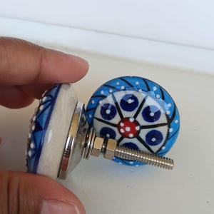 Peut inclure: Deux boutons de placard ronds en c&eacute;ramique avec un motif peint &agrave; la main, un point rouge central entour&eacute; de cercles bleu fonc&eacute;, de lignes noires et de points blancs sur fond bleu clair. Chaque bouton a un boulon et un &eacute;crou en m&eacute;tal argent&eacute;.