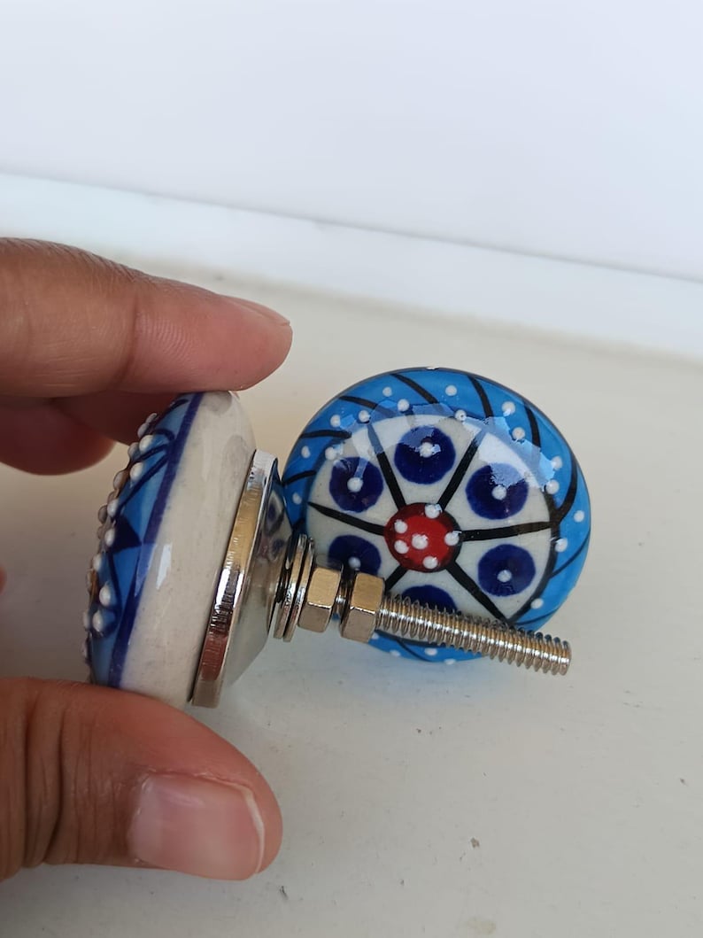 Peut inclure: Deux boutons de placard ronds en c&eacute;ramique avec un motif peint &agrave; la main, un point rouge central entour&eacute; de cercles bleu fonc&eacute;, de lignes noires et de points blancs sur fond bleu clair. Chaque bouton a un boulon et un &eacute;crou en m&eacute;tal argent&eacute;.