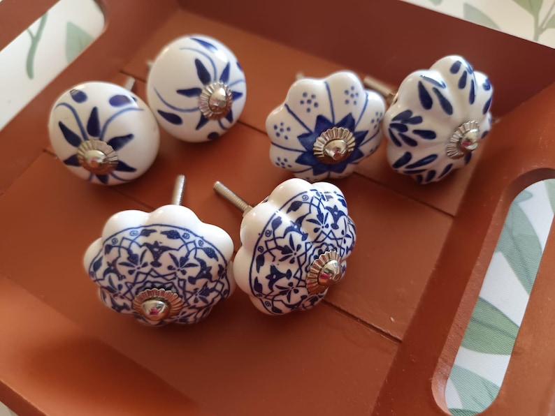 Peut inclure: Un ensemble de six boutons de placard en c&eacute;ramique pr&eacute;sentant des surfaces blanches avec des motifs peints en bleu. Les boutons comprennent deux formes rondes lisses et quatre designs &agrave; bords festonn&eacute;s, chacun avec une fixation m&eacute;tallique argent&eacute;e.