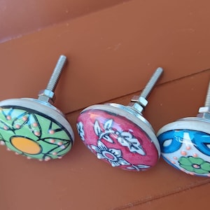Peut inclure: Trois boutons de placard ronds en c&eacute;ramique avec quincaillerie m&eacute;tallique. Le bouton de gauche est vert avec un centre jaune et des points orange. Le bouton central est rose avec des motifs floraux blancs. Le bouton droit est bleu avec une fleur verte et des points orange.