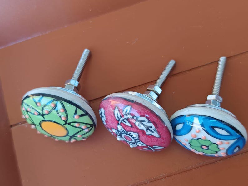 Peut inclure: Trois boutons de placard ronds en c&eacute;ramique avec quincaillerie m&eacute;tallique. Le bouton de gauche est vert avec un centre jaune et des points orange. Le bouton central est rose avec des motifs floraux blancs. Le bouton droit est bleu avec une fleur verte et des points orange.