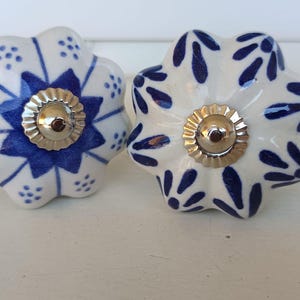 Peut inclure: Deux boutons de tiroir ronds en c&eacute;ramique avec un bord festonn&eacute;. Le bouton de gauche pr&eacute;sente un motif d'&eacute;toile bleue avec de petits points, tandis que celui de droite a un motif de feuilles bleues. Les deux ont un capuchon central argent&eacute;.