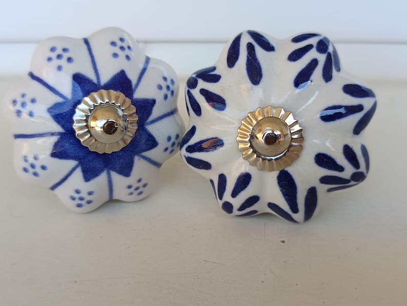 Peut inclure: Deux boutons de tiroir ronds en c&eacute;ramique avec un bord festonn&eacute;. Le bouton de gauche pr&eacute;sente un motif d'&eacute;toile bleue avec de petits points, tandis que celui de droite a un motif de feuilles bleues. Les deux ont un capuchon central argent&eacute;.