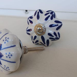 Peut inclure: Deux boutons de tiroir en c&eacute;ramique avec une base blanche et un motif floral bleu. Chaque bouton a une forme festonn&eacute;e rappelant une citrouille et un capuchon central argent&eacute;. Une tige filet&eacute;e m&eacute;tallique d&eacute;passe de l'un des boutons.