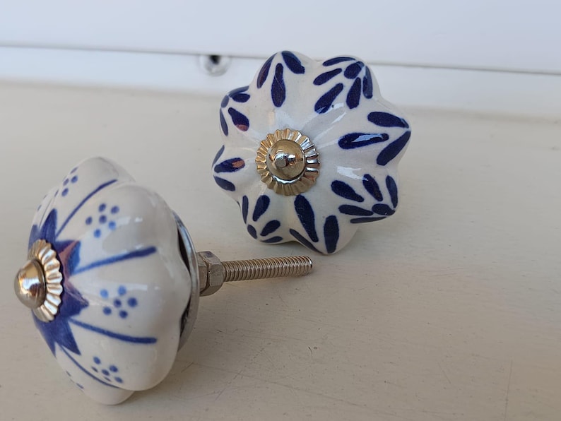Peut inclure: Deux boutons de tiroir en c&eacute;ramique avec une base blanche et un motif floral bleu. Chaque bouton a une forme festonn&eacute;e rappelant une citrouille et un capuchon central argent&eacute;. Une tige filet&eacute;e m&eacute;tallique d&eacute;passe de l'un des boutons.
