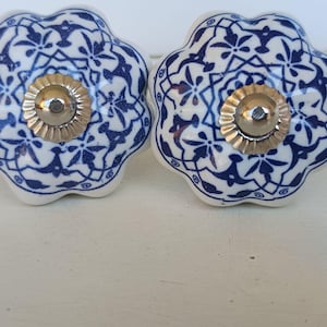 Peut inclure: Deux boutons en c&eacute;ramique en forme de fleur avec une base blanche et un motif g&eacute;om&eacute;trique et floral bleu fonc&eacute; complexe. Chaque bouton poss&egrave;de un capuchon central m&eacute;tallique argent&eacute; cannel&eacute; avec une petite t&ecirc;te de vis visible au centre.