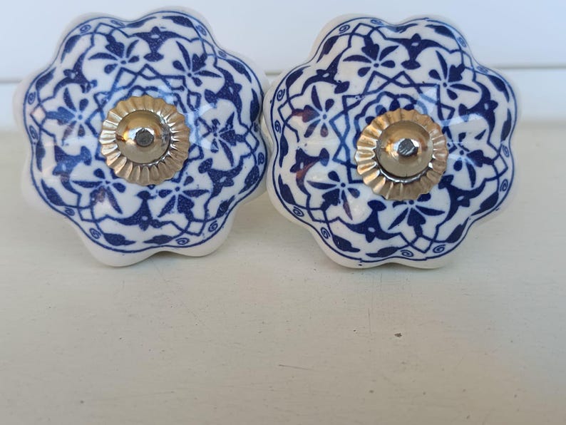 Peut inclure: Deux boutons en c&eacute;ramique en forme de fleur avec une base blanche et un motif g&eacute;om&eacute;trique et floral bleu fonc&eacute; complexe. Chaque bouton poss&egrave;de un capuchon central m&eacute;tallique argent&eacute; cannel&eacute; avec une petite t&ecirc;te de vis visible au centre.