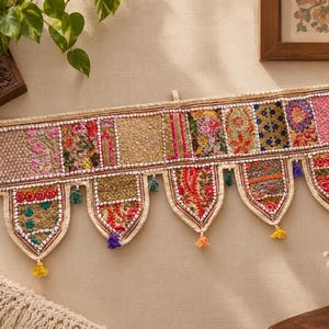 May include: A rectangular wall hanging featuring a patchwork of embroidered fabric panels in various colours including pink, gold, green, and blue. The bottom edge has five pointed, downward-hanging sections, each finished with small colourful tassels.