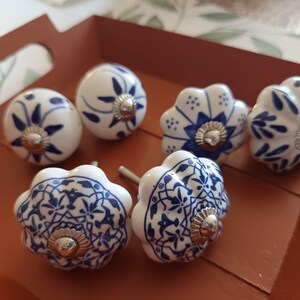 Peut inclure: Un ensemble de six boutons de placard en c&eacute;ramique pr&eacute;sentant des surfaces blanches avec des motifs floraux et g&eacute;om&eacute;triques bleus complexes. Chaque bouton a une forme arrondie ou festonn&eacute;e et inclut une fixation m&eacute;tallique argent&eacute;e.