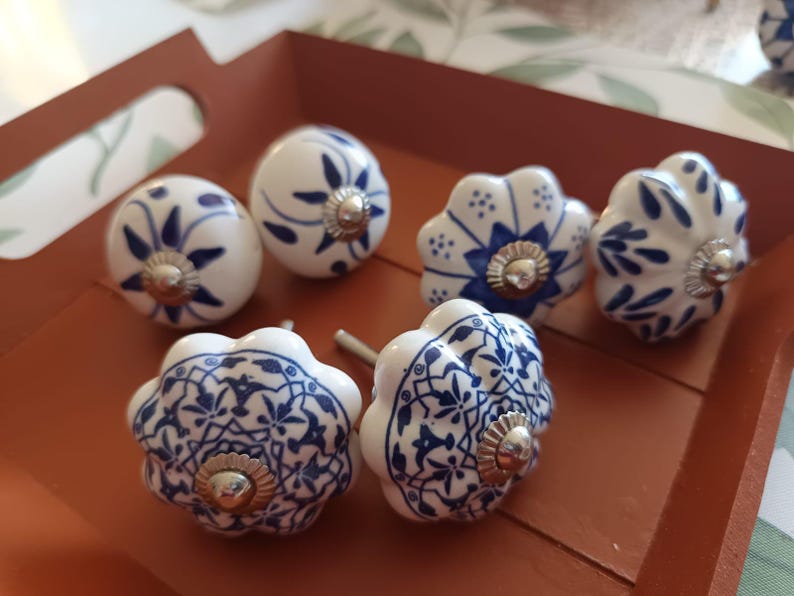 Peut inclure: Un ensemble de six boutons de placard en c&eacute;ramique pr&eacute;sentant des surfaces blanches avec des motifs floraux et g&eacute;om&eacute;triques bleus complexes. Chaque bouton a une forme arrondie ou festonn&eacute;e et inclut une fixation m&eacute;tallique argent&eacute;e.